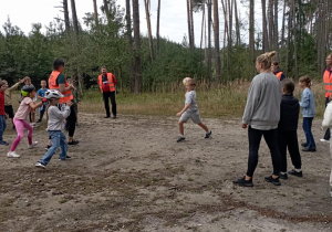 Dzieci z klasy 2b i opiekunowie grają w piłkę na leśnej polanie. Wszyscy uczestnicy są uśmiechnięci, a część dzieci nosi kaski rowerowe.