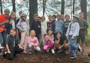 Klasa 2b z wychowawczynią Moniką Mettler podczas wycieczki rowerowej w lesie, przed napisem „2B” ułożonym z szyszek. Dzieci pozują do wspólnego zdjęcia.