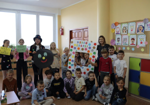 Klasa 1b z wychowawczynią Agnieszką Jarzębowską podczas Dnia Kropki. Uczniowie pozują do wspólnego zdjęcia z kolorowymi kropkami na ubraniach. W tle widać dekoracje z pracami plastycznymi i plakatem „Dzień Kropki”.