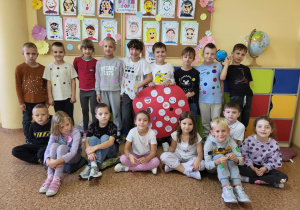 Uczniowie klasy 1b Szkoły Podstawowej nr 13 w Pabianicach pozują wspólnie podczas obchodów Dnia Kropki. Dzieci ubrane są w stroje z kolorowymi kropkami. Na środku znajduje się czerwony plakat – „Kropka Mocy” z rysunkami i hasłami. W tle widać galerię portretów i kolorową dekorację.