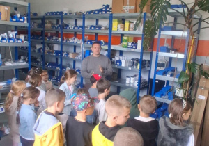 Uczniowie klasy 2B słuchają pracownika MZK, który prezentuje magazyn z częściami do autobusów. Dzieci stoją wśród regałów z uporządkowanymi elementami technicznymi.