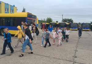 Dzieci z klasy 2b SP13 idą przez zajezdnię MZK wśród żółto-niebieskich autobusów, w tle budynek i opiekunowie.