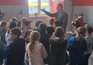 Dzieci z klasy 2b słuchają mężczyzny opowiadającego o pracy wulkanizatora; w tle maszyny warsztatowe i okno z doniczkami na parapecie.