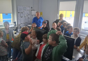 Grupa dzieci z klasy 2b w pomieszczeniu dyspozytorni MZK, słuchają pracownika, wokół stanowiska komputerowe i dokumentacja na ścianach.