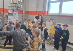 Grupa dzieci z klasy 2b w hali technicznej MZK słucha pracownika prezentującego elementy autobusu; w tle nauczyciele i stanowisko z dokumentami.