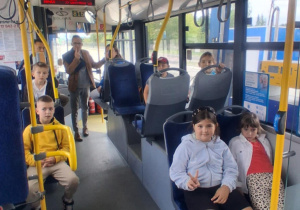 Uczniowie klasy 2B SP13 siedzą w miejskim autobusie Solaris podczas wycieczki do MZK. Uśmiechnięci, w towarzystwie opiekuna, pokazują znak pokoju do aparatu.