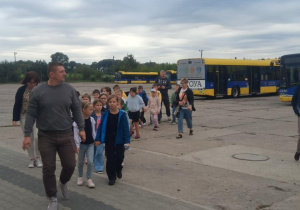 Uczniowie klasy 2B SP13 z nauczycielkami idą w parach po terenie MZK w Pabianicach. W tle widoczne są autobusy w barwach żółto‑niebieskich. Trwa edukacyjna wycieczka.