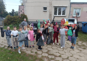 Grupa dzieci z kolorowymi workami na śmieci stojąca przed szkołą po zakończonej akcji sprzątania, w tle kontenery i pojemnik na odzież.