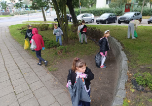 Grupa uczniów sprzątająca teren zielony przy ulicy – dzieci w rękawiczkach trzymają worki na śmieci i zbierają odpady z trawnika i krzewów.
