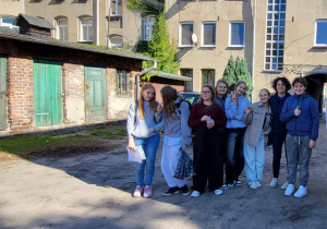 Pani Sylwia Muszyńska i uczniowie klasy 8a stoją na podwórku przy kamienicy z zielonymi drzwiami komórek. W tle jasna elewacja i słońce rzucające cienie drzew.