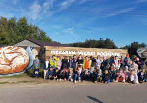 Grupa uczniów przed ogrodzeniem z napisem „Piekarnia Wiejski Bochenek”, obok namalowane bochenki chleba, w tle las i niebieskie niebo.