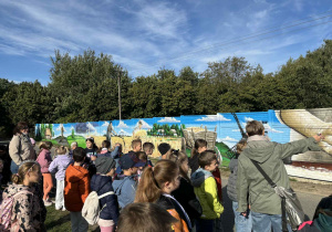 Uczniowie klasy 2A i 2B oglądają mural o tematyce wiejskiej, a nauczycielka Monika Mettler wskazuje fragment malowidła. W tle drzewa i błękitne niebo.