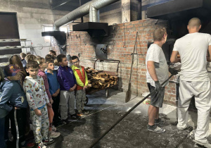Uczniowie klasy 2A i 2B obserwują piekarzy wkładających chleb do pieca opalanego drewnem wewnątrz tradycyjnej piekarni.