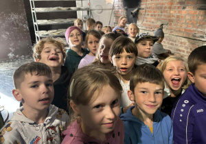 Uczniowie klasy 2A i 2B w piekarni, uśmiechnięci stoją w grupie na tle ceglanej ściany i pieca chlebowego.