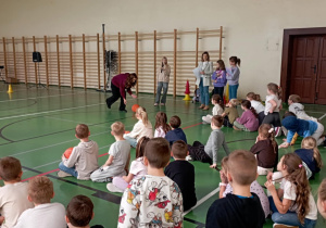 Dzieci siedzące na sali gimnastycznej, kibicujące podczas wyścigu z piłką w ramach Dnia Integracji. Na boisku uczennica biegnie z piłką, a prowadzące i nauczycielki obserwują przebieg konkurencji.