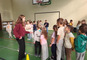Dzieci z klas I–III w kolejce do zabawy podczas Dnia Integracji w sali gimnastycznej. Starsza uczennica prowadzi zajęcia z mikrofonem.