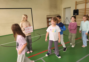 Dziewczynka trzymająca tabliczkę „Stacja uśmiechów” podczas zabawy integracyjnej; uczniowie przechodzą obok niej z uśmiechami – scena z Dnia Integracji w sali gimnastycznej.