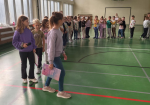 Dzieci ustawione w parach na sali gimnastycznej podczas Dnia Integracji; dwie prowadzące dziewczynki idą na czele z notatnikami.