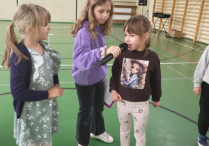 Trzy dziewczynki uczestniczą w Dniu Integracji – jedna mówi do mikrofonu trzymanego przez koleżankę, trzecia dziewczynka uśmiecha się obok.