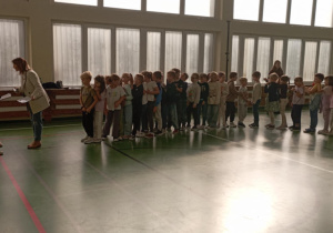 zieci stojące w dwóch rzędach na sali gimnastycznej tworzą „Pociąg przyjaźni”; z przodu uczennice i nauczycielka koordynują przebieg wydarzenia.