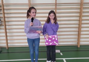 Dwie uczennice prowadzące wydarzenie szkolne stojące przy drabinkach gimnastycznych; jedna z nich mówi do mikrofonu.