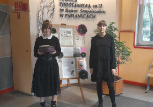 Dwie uczennice w czarnych strojach stoją w holu szkoły przy gazetce poświęconej patronce Stefanii Sempołowskiej. Jedna trzyma kosz z cukierkami, obok widoczne są prace plastyczne.