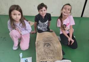 Troje dzieci siedzi na sali gimnastycznej obok dużego arkusza papieru z rysunkiem postaci i napisem „KUKAIGAHLA”. Uśmiechnięte pozują do zdjęcia po zakończonej pracy plastycznej.