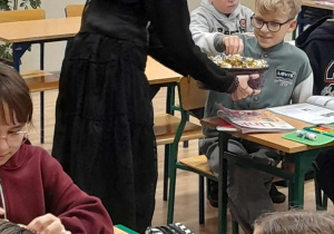 Uczennica w czarnej sukience rozdaje cukierki uczniom podczas lekcji języka angielskiego. Dzieci siedzą przy stolikach z otwartymi podręcznikami i piórnikami.