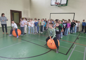 Uczniowie klas I–III podczas zawodów w sali gimnastycznej. Dwoje dzieci skacze na dużych pomarańczowych piłkach z buzią kota, a reszta uczniów stoi w rzędach i kibicuje.