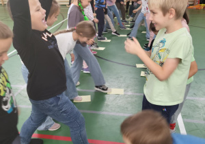 Uczniowie klas młodszych bawią się w sali gimnastycznej. Stoją w dwóch rzędach i wykonują zadanie z kartkami na podłodze. Jedno z dzieci unosi ręce w górę w geście radości.