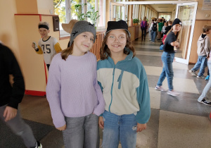 Dwie uczennice stoją na szkolnym korytarzu podczas „Dnia kapelusza”. Jedna ma na głowie szarą czapkę zimową, druga czarny kapelusz typu bucket i okulary. W tle widać innych uczniów uczestniczących w wydarzeniu.