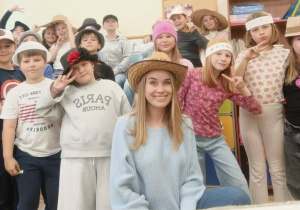 Grupa uczniów klasy 3 pozuje w sali lekcyjnej razem z wychowawczynią. Wszyscy mają na głowach różnorodne kapelusze i fantazyjne nakrycia głowy — słomkowe, materiałowe i papierowe — w ramach „Dnia kapelusza”. Wychowawczyni siedzi na pierwszym planie w kapeluszu słomkowym.