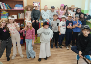 Grupa uczniów klasy 2 pozuje w sali lekcyjnej podczas „Dnia kapelusza”. Dzieci mają na głowach różnorodne nakrycia — słomkowe kapelusze, papierowe korony, czapki sportowe i fantazyjne projekty wykonane własnoręcznie. Wszyscy stoją lub siedzą, pozując do wspólnego zdjęcia.