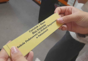 Zbliżenie dłoni dziecka trzymającego imienną żółtą zakładkę do książki z napisem „Czytelnik I Klasa! Maja Krajewska. Szkoła Podstawowa im. Stefanii Sempołowskiej w Pabianicach”. W tle widoczne rozmyte sylwetki innych uczniów.