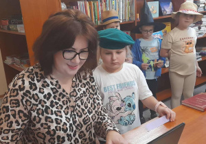 Nauczyciel bibliotekarz siedzi przy biurku i wpisuje dane ucznia do komputera podczas pasowania pierwszoklasistów na czytelników. Obok stoi dziewczynka w zielonym beretowym kapeluszu, a w tle widać inne dzieci w kapeluszach czekające na swoją kolej.