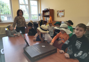 Uczniowie klasy 1 siedzą przy stole w bibliotece szkolnej, słuchając nauczyciela bibliotekarza podczas pasowania na czytelników. Dzieci mają na głowach różnorodne kapelusze, a na stole leży duży „Atlas Rzeczypospolitej Polskiej”.