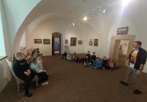 Grupa uczniów siedzących w sali muzealnej w Pabianicach, oglądających wystawę i słuchających prowadzącego zajęcia.