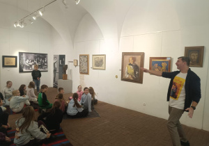 Uczniowie siedzący w sali muzeum w Pabianicach, słuchający przewodnika omawiającego obrazy wiszące na ścianie.