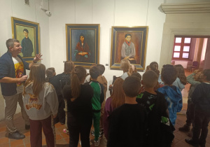 Grupa uczniów stojących przed trzema portretami w muzeum, słuchających przewodnika opowiadającego o obrazach.