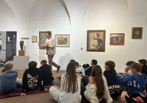 Przewodnik opowiada uczniom siedzącym na podłodze o obrazach i rzeźbach na wystawie Kislinga w Pabianicach.