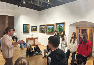 Przewodnik rozmawia z uczniami na tle pejzażowych obrazów Kislinga; młodzież słucha w skupieniu w sali muzeum.