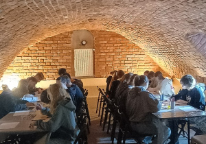 Uczniowie klasy 8b siedzą przy stołach w ceglanej sali muzeum i wykonują prace plastyczne podczas warsztatów.