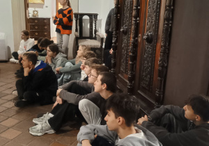 Uczniowie klasy 8b siedzą na podłodze w muzeum i słuchają przewodnika w sali z zabytkowymi meblami i obrazami.