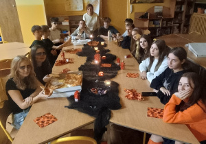 Grupa uczniów klasy 8b siedzi przy dużym stole w szkolnej świetlicy, udekorowanej w halloweenowym stylu. Na stole stoją zapalone świeczki i jedzenie, a uczniowie jedzą i rozmawiają w przyciemnionym nastrojowym oświetleniu.