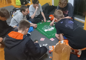 Grupa uczniów klasy 8b siedzi na podłodze w sali szkolnej i gra w karty, korzystając z maty do gier i żetonów. Obok nich stoją napoje i przekąski. Uczniowie spędzają czas na integracji podczas nocnego wydarzenia w szkole.