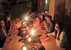 Kilka uczennic klasy 8b siedzi przy długim stole w ciemnej sali, oświetlonej jedynie świecami. Na stole znajdują się halloweenowe dekoracje, przekąski i napoje. Widoczne są motywy pajęczyn i jesienne światła, tworzące klimatyczną atmosferę nocnego wydarzenia w szkole.