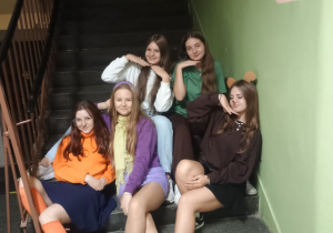 Pięć uczennic klasy 8b siedzi na schodach w szkole podczas nocnej imprezy. Dziewczyny pozują uśmiechnięte, część trzyma dłonie pod brodą w humorystycznym geście. W tle widać poręcz schodów i zieloną ścianę.