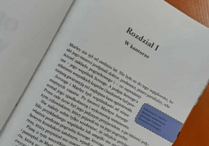 Otwarta książka na blacie, widoczny tytuł rozdziału „Rozdział I. W kantorze” z „Opowieści wigilijnej”; obok leży tablet.