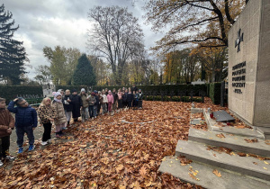 Uczniowie stojący w rzędzie przy pomniku poległych żołnierzy na cmentarzu jesienią, otoczeni opadłymi liśćmi i krzyżami nagrobnymi w tle. Atmosfera pamięci i refleksji.