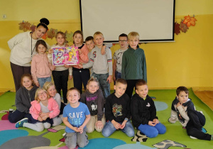 Grupa uczniów świetlicy szkolnej z nauczycielką prezentuje ułożone puzzle po zakończeniu zabawy integracyjnej. Dzieci stoją i siedzą na kolorowej macie, uśmiechając się do zdjęcia.